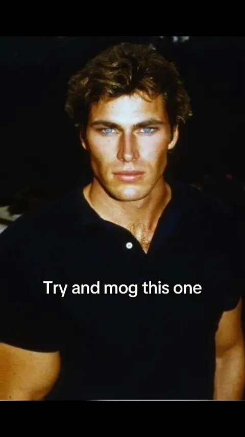 A portrait of a man with slicked-back, wavy brown hair and striking blue eyes. He wears a black collared shirt and has a slight smirk. The background is a dark, out-of-focus area.