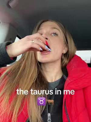 A woman in a car with long, light brown hair is the central focus, her face slightly angled towards the viewer. She has pale skin, blue eyes, and red and blue painted nails. Her lips are slightly parted, showing a hint of blue lip color, and she's holding a finger up near her mouth. She wears a red puffy jacket.
