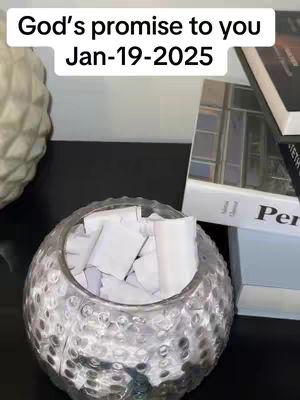 The image shows a clear, close-up shot of a decorative glass bowl filled with white, rolled-up paper notes. The bowl is in the foreground, slightly off-center, with a blurred, textured surface. Behind it, out of focus, is a pile of books and other decorative objects on a dark surface. A text overlay at the top reads 'God's promise to you' and 'Jan-19-2025'. The lighting is soft and diffused, enhancing the reflective qualities of the glass bowl and the neutral color palette.