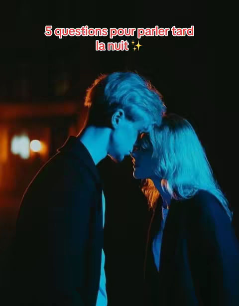 A couple is captured in a close embrace at night. The man, on the left, has light-colored hair and wears a dark coat, while the woman, to his right, has wavy blonde hair and a similar dark top. Both are seen in profile, gazing at each other with their foreheads touching. The lighting is dramatic, the backdrop is dark, possibly an urban night scene, with a blurred orange glow. The overall mood is intimate and romantic.