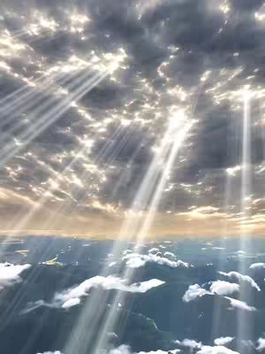 The image captures a serene, aerial view of clouds bathed in sunlight, with rays of light piercing through the cloud cover. The composition emphasizes the interplay of light and shadow, creating a dramatic and heavenly atmosphere. The color palette ranges from bright whites and golden hues in the illuminated areas to darker blues and grays within the clouds and in the distant landscape.