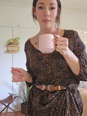 A woman holds a pink polka-dotted mug, appearing to pose in front of a light-colored wall with a plant and a small wooden shelf. She is wearing a patterned dress with a belt. A bed, wooden stool, and potted plant also appear in the background.