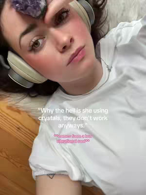 A selfie shows a young woman lying down, gazing towards the camera. She's wearing headphones and has a purple crystal resting on her forehead. Her cheeks are flushed with pink blush, and she wears light makeup. Text is overlaid, questioning her use of crystals. The background appears to be a light-colored fluffy surface and wooden floor.