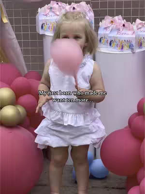 A young girl is holding a large pink cotton candy in front of her face. She is wearing a white dress and stands surrounded by large pink balloons and party decorations. The image is brightly lit, with a focus on the girl and the cotton candy.