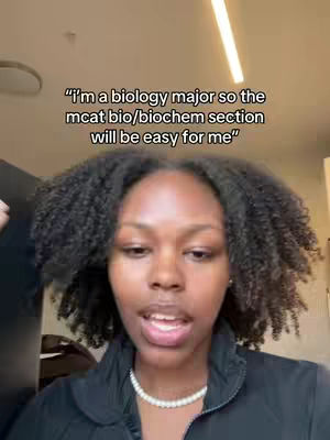 A young woman with dark curly hair is centered in the frame, looking directly at the camera and speaking. She wears a black jacket and a pearl necklace. The background is blurred, suggesting an indoor setting with a glimpse of a ceiling and a light fixture. Text overlayed on the image reads, "I'm a biology major so the mcat bio/biochem section will be easy for me". The overall composition is a medium shot with a focus on the subject's face and upper body.
