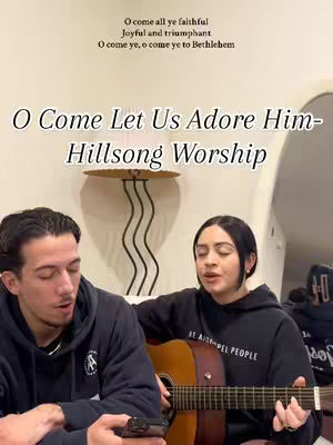 A cover image shows a man on the left and a woman on the right, both positioned from the chest up with a neutral indoor background. The man is holding a smartphone while looking at the woman. The woman is sitting and playing an acoustic guitar. Text overlays stating "O Come Let Us Adore Him - Hillsong Worship".