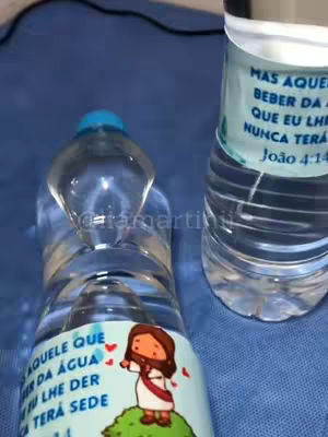 The image shows two partially visible water bottles on a blue surface. The bottle on the left has a label with an illustration of a cartoon Jesus figure standing on a green patch with text about the water of life. The bottle on the right has a text label with a Bible verse. The image is lit with soft, natural light.