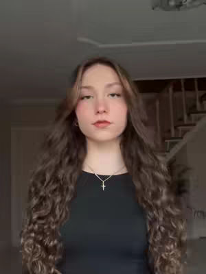 A young woman with long, wavy brown hair, wearing a black top and a silver cross necklace, poses indoors with a neutral expression. The background includes a staircase and a glimpse of a ceiling.