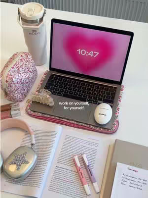 A still life featuring a laptop displaying a pink heart shape with the time 10:47. Surrounding the laptop are study essentials: open books with handwritten notes, highlighters, a water bottle, a makeup bag, headphones, and a Chanel-branded item.