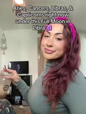 A woman with long, reddish-purple hair wearing a headband gazes slightly off-camera with a subtle smile. The background is a well-lit living space with a television, and a drink is held in her hand. Text overlays at the top of the image read: Aries, Cancers, Libras, & Capricorns right now under this Full Moon in Libra.