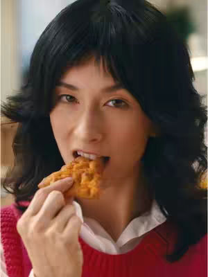 A close-up shot of a woman with long, dark hair and light skin, biting into a piece of pita chip. She is wearing a red sweater over a white collared shirt. The chips are golden brown, and the background is softly blurred.