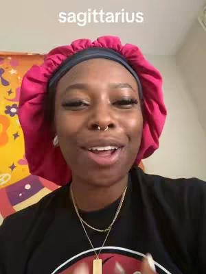 A person with dark skin and hair is the primary focus of the image. She has a pink satin head covering, a small nose ring and a necklace. The background is a room with a yellow decorative wall.
