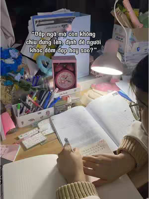 A person is studying at a desk filled with stationery and study materials. The person is sitting at a desk with an open book and a pen in hand, writing on a sticky note. There's a pink alarm clock in a box and a lamp on the desk. The overall setting suggests a study environment.