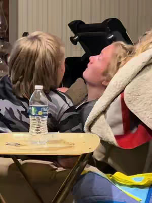 The image shows two people in a close-up scene, likely indoors. On the left, a person with short, light-colored hair is partially visible from the back, wearing a camouflage-patterned top. They are facing the other person in the image. To the right is a person with their head back, eyes closed and blonde hair, who is resting against a beige and red blanket. A water bottle sits atop a small, light-brown table in the foreground, slightly obscuring the view. The setting suggests an intimate space, with neutral-colored walls and indirect lighting.
