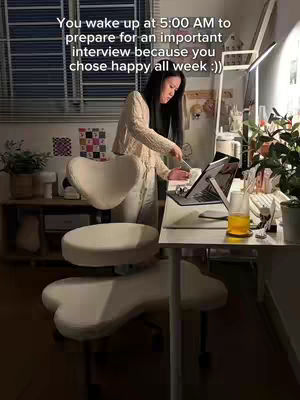 A young woman is standing in her home office, preparing for an interview. She's dressed in a cream-colored sweater and pants, with long dark hair. The room is well-lit with a desk, chair, and other office supplies. The background includes a window with blinds, plants, and personal items, creating a cozy and focused workspace.