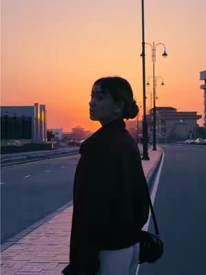 A woman stands on a city street at sunset, silhouetted against the vibrant orange and pink sky. She is facing left, with her body turned slightly away from the camera, her head is angled towards the camera. Beside her are streetlights, and a road in the background.
