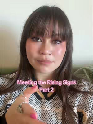 A woman with long, dark hair and bangs looks towards the camera. Text overlayed on the image says 'Meeting the Rising Signs Part 2' in a pink font. She is wearing a mesh top and has a hand up with a finger out, showing off a ring.