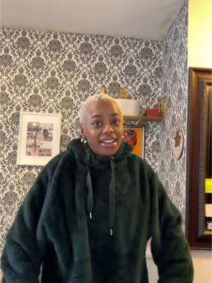 A woman with short, light-colored hair is centered in the frame, wearing a dark green fuzzy hoodie. Her expression is friendly and she appears to be speaking. The background includes a patterned wallpaper and framed artwork.