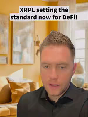 A man in a black shirt is centered in the frame, appearing to be in a living room setting. Overlaid text in a white rectangular box reads, "XRPL setting the standard now for DeFi!" The background includes a blurred-out window, artwork, and a portion of a couch. The color palette is warm, with yellows and creams dominating, with the man's skin contrasting with the text and background.