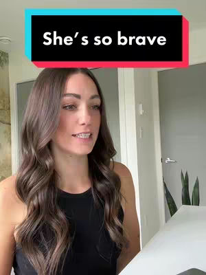 A woman with long brown hair is positioned in front of a light-colored setting with a sign above her that says 'She's so brave'. She has a black top on and is looking at the camera. The background has a minimalist appearance.