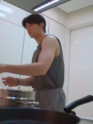A man wearing a gray tank top and sweatpants is standing in a kitchen, looking to his left while cooking with a pot in the middle. The background features white cabinets and walls, with a spotlight.