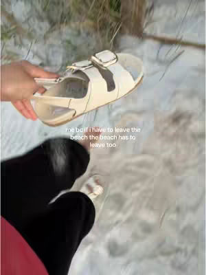A person's lower body and arm are visible, holding a cream-colored sandal in front of a sandy beach background. The sandal is a closed-toe design with a buckle. The sand appears white with some green brush peeking through. Text overlays the image.