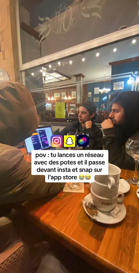 The cover image shows three young men sitting around a wooden table in what seems to be a cafe or restaurant. A laptop is open on the table, and the men appear to be engaged in a conversation or activity related to it. The setting includes the interior of a cafe, suggested by the visible decor and the presence of empty coffee cups and a wine glass. The lighting is a mix of indoor illumination and possible natural light from a window. Three social media logos are layered with the text. The overall composition indicates a casual, social setting likely used for discussing media and app development.