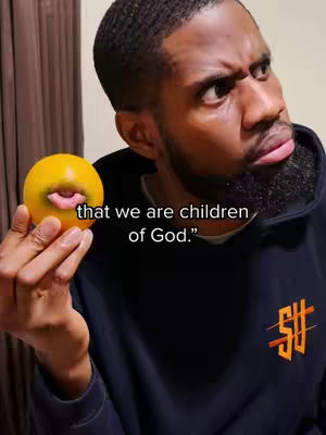 A man with a beard and a serious expression holds a yellow orange with a mouth and tongue drawn on, like a mouth that's talking. The man is wearing a black hoodie with a logo in orange color. The background appears to be a simple interior setting, the text on the image says "that we are children of God."