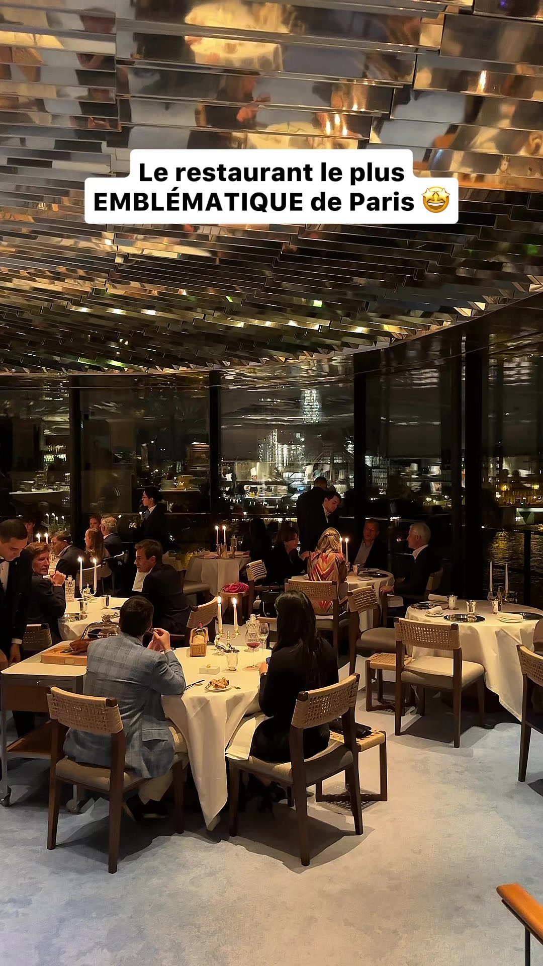 An interior shot of a high-end restaurant in Paris at night. The ceiling is covered in mirrored panels. Tables are set with white tablecloths, candles, and glassware. Diners are seated around the tables, some facing the camera, others with their backs turned. The background includes large windows offering a view of the city lights. The lighting is dim, giving a warm and elegant feel. Guests are dressed in semi-formal attire. The overall composition conveys a sense of luxury and dining experience.