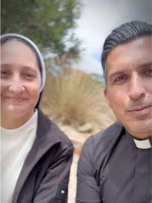 Two people are in a selfie-style shot. On the left is a woman wearing a black jacket over a sweater and white headcovering. Beside her is a man wearing a black shirt and a clerical collar, with a clear backdrop of trees and brush. The composition is a close-up.