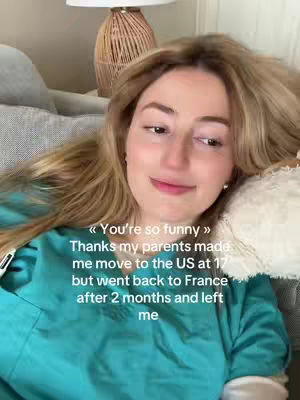A young woman with long blonde hair is seen reclining on a couch with light-colored pillows. She is wearing a turquoise scrub top. Text overlayed shows a quote from her parents' making her move to the US at 17, but then moving back to France, which prompts a facial expression of thought and slight amusement.