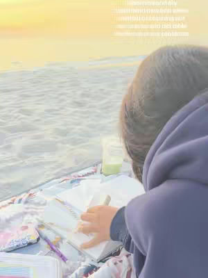 A person in a blue hoodie sits on a blanket by the water, writing in a notebook. The person's back is to the viewer. The background is a soft yellow and the water is a hazy grey.