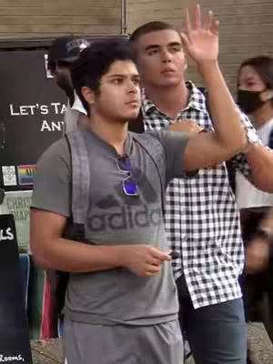 A medium shot features two young men standing close together outdoors. The main subject, on the left, is wearing a gray t-shirt with the Adidas logo, a gray backpack, and sunglasses hanging from his shirt. He's looking towards the side with a serious expression. The man on the right, slightly taller, is raising his uncovered hand as to wave, with a checkered shirt and dark pants; he's looking upwards. Some individuals in the background are blurred, one wearing a black cap reading something. The overall scene appears to be a public gathering or event with religious overtones, suggested by the backdrop.