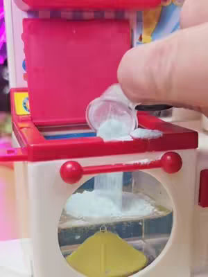 A close-up shot of a vintage toy playset. A hand is pouring light blue granules from a small transparent plastic cup into the playset. The playset is mostly white, with red and pink accents. The background is blurred and includes hints of purple.