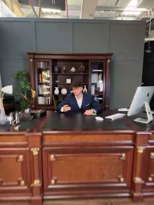 A man is seated behind a large, ornate wooden desk, with a dark leather top. He is wearing a dark suit and white shirt. Behind him is a large wooden cabinet with glass-fronted doors filled with books and decorations. The background consists of a dark gray wall and a window. The overall lighting suggests an office setting, and the composition is framed to show the subject engaging with what appears to be a phone or device.