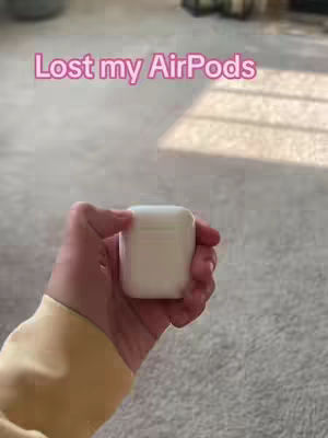 A hand holding the white charging case for a pair of Apple AirPods. The case is centered in the frame, with the person's hand and part of their yellow sleeve visible. The background is a light-colored carpet. Text overlayed in pink reads 'Lost my AirPods'.