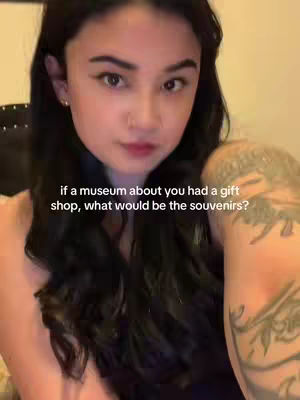 A young woman with long, dark hair is centered in the frame, gazing towards the camera. She has a delicate facial structure and is wearing minimal makeup. The background is softly illuminated, and a portion of her arm, adorned with a tattoo, is visible on the right side of the image. Text overlay is present on the bottom half of the image.