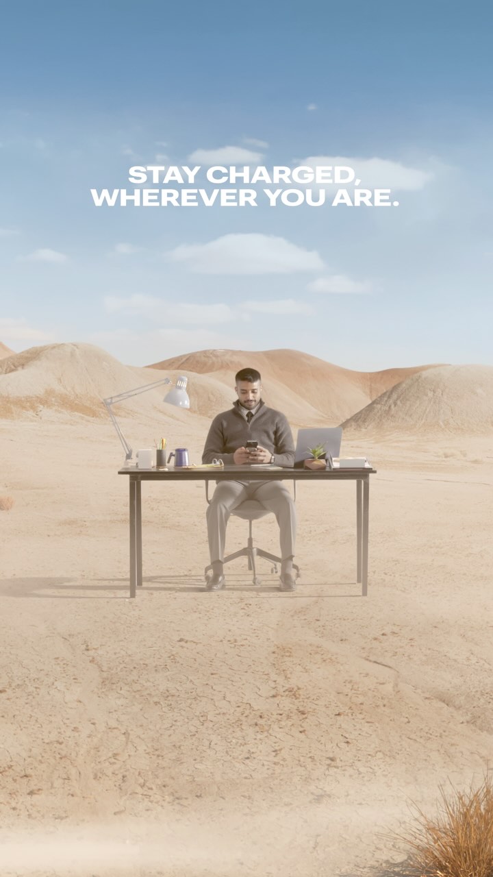 A man sits at a desk in a desert landscape. Text overlay says "STAY CHARGED, WHEREVER YOU ARE."