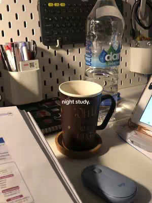 A well-lit desk setup features a brown mug on a wooden coaster, a half-full water bottle, a keyboard, and a journal. The image is predominantly neutral tones, with the mug and notebook being the focal points. The lighting suggests a nighttime study environment.