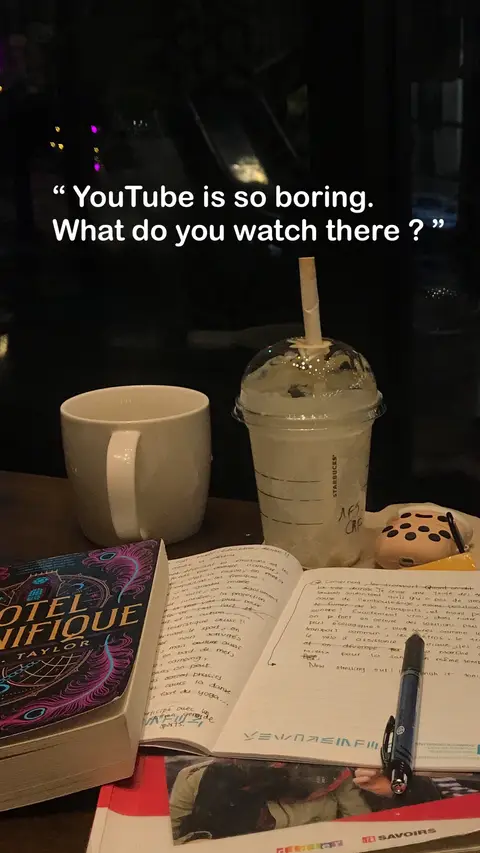 The image shows a table setting with a white coffee mug to the left, a book, an open notebook with handwritten notes, a drink in a Starbucks cup with a straw, and a pastry. The background is dark, suggesting a low-light environment.