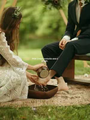 A woman in a white dress is washing the feet of a man sitting on a wooden bench. The scene is set outdoors with green foliage in the background. The woman is pouring water from a small brown jug into a wooden bowl where the mans feet are. Text overlayed onto the image says, 'POV: O livro preferido dela é ESTER.'