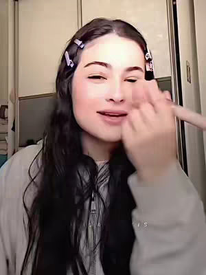 A selfie-style shot shows a young woman with long, dark hair, parted on the side, and held back with bobby pins. She has fair skin, with blush applied to her cheeks. She is in the process of applying makeup, the brush is visible in her right hand near her face, with her eyes closed. The background is a simple, off-white or cream-colored wall, and a hint of a closet or wardrobe is visible on the right side.