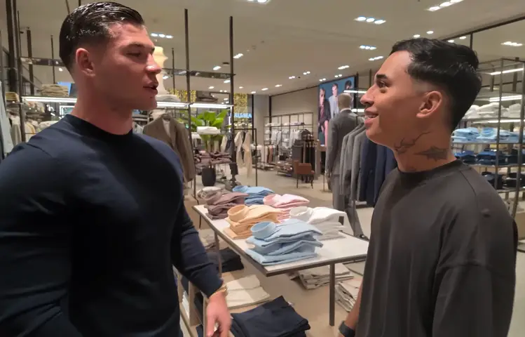Two men are talking inside a clothing store. One man, on the left, is fair-skinned with short, slicked-back hair and is wearing a dark blue shirt. The other man on the right has dark hair and visible neck tattoos and is wearing a gray t-shirt. The background includes clothing racks and store displays.
