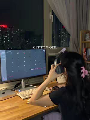 A person with dark hair, wearing a black shirt and pink hair clip, is sitting at a desk at night, with a computer and headphones, looking into the monitor. A large window is a backdrop showing city lights.