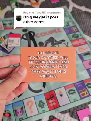 A hand holds an orange card with text over a board game setting. The game board is primarily blue and depicts the word COUPLE on it. A comment bubble with text is at the top left corner of the image. The card has text on it that says: GO TO JAIL "YOUR PARTNER WILL TIE YOUR HANDS AND CAN DO WHATEVER THEY WANT FOR 2 MINUTES."