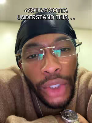 A close-up shot of a Black man with a durag, glasses, and a wristwatch. He is looking directly at the camera, his lips slightly parted as if speaking. The background is uncluttered, focusing attention on the subject.