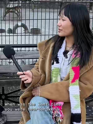 A woman with long black hair sits on a bench in front of a black metal fence, holding a microphone. She is wearing a brown coat, a white shirt, and a colorful scarf. The background features trees and a view of a public park.