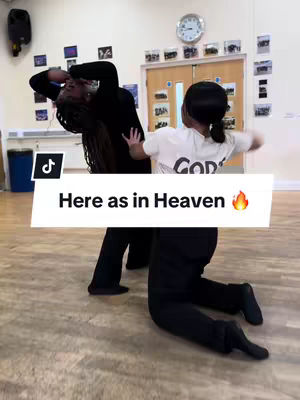 The image captures a dance studio scene with two dancers in action. One dancer, wearing a white t-shirt and black shorts, is in a dynamic pose with arms extended. The other dancer, dressed in all black, is kneeling, with one arm raised. The setting is a dance studio lit by overhead lights, and the floor is a light wood color.