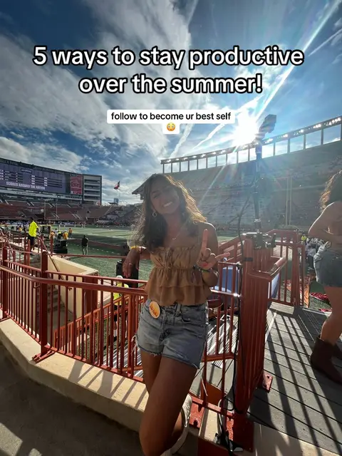 A woman in a tan top and denim shorts gives a peace sign while standing in front of a stadium setting with a cloudy sky.
