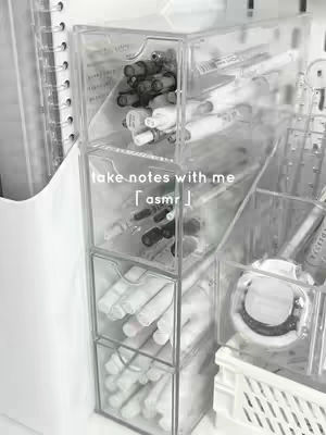 A close-up shot of a clear, multi-compartment organizer filled with pens, markers, and other stationery items. The organizer is the focal point, while items like a notebook and some other containers are blurred in the background. The image has a clean, minimalist aesthetic, with a focus on neatness and organization, enhanced by a black and white color scheme.